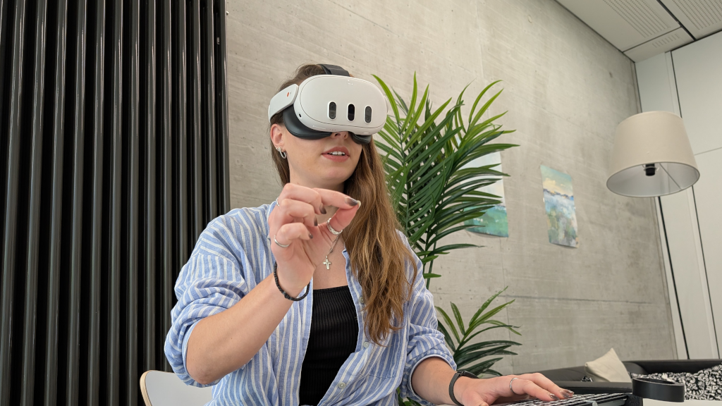 Foto: Frau mit VR-Brille macht eine Kneifgeste mit der rechten Hand, während sie mit der linken die Tastatur bedient.